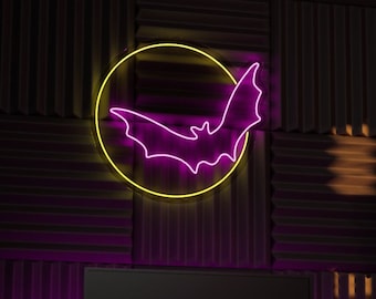 May include: A neon sign featuring a bat inside a yellow circle is mounted on a dark wall. The room has a desk with a computer monitor, microphone, and speakers. The wall is made of dark acoustic panels and brick.