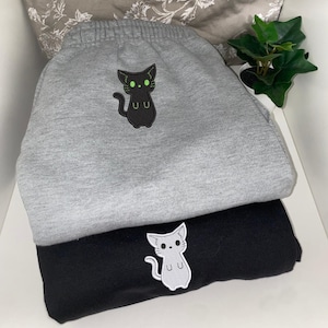 Może przedstawiać: Dwie pary spodni dresowych z wzorem kota. Górna para jest szara z czarnym kotem o zielonych oczach i tekstem "UU". Dolna para jest czarna z szarym kotem o zielonych oczach i tekstem "UU".