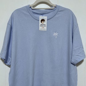 May include: A light blue t-shirt with a small embroidered palm tree on the front. The t-shirt is hanging on a hanger. The tag on the t-shirt says "Embroidered Apparel & Gifts".