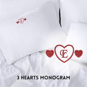 May include: White pillowcases with red embroidered hearts and a monogram letter. The text "3 HEARTS MONOGRAM" is displayed at the bottom of the image. The bedding is arranged on a white bedspread.