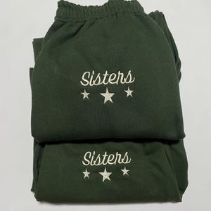 Może przedstawiać: Dwie pary ciemnozielonych spodni dresowych ułożone jedna na drugiej. Na każdej parze wyhaftowano białą czcionką słowo "Sisters", pod którym znajdują się trzy białe gwiazdki. Spodnie dresowe mają elastyczne ściągacze w pasie.