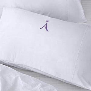 May include: White pillowcase with a purple embroidered letter "A" and a crown above it. The pillowcase is made of a smooth, white fabric and has a sewn seam along the edge. The pillowcase is part of a bedding set.