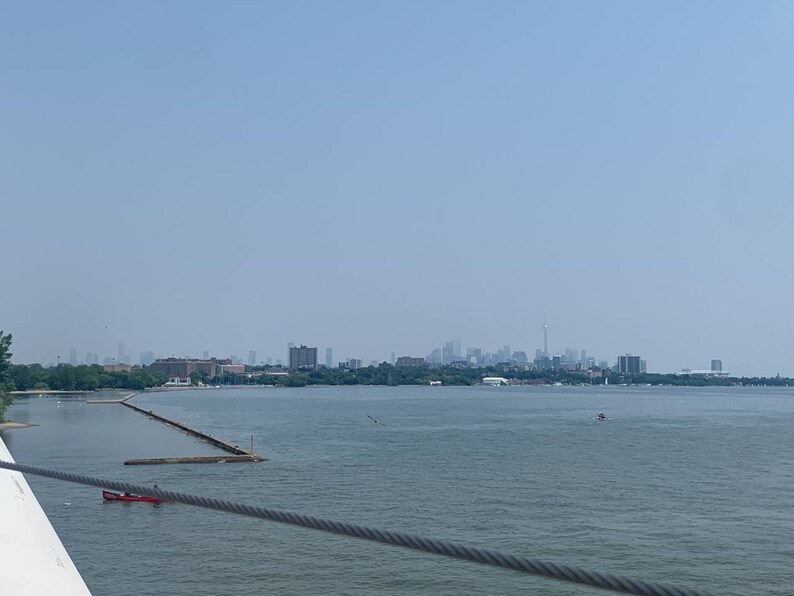 May include: A scenic view of a city skyline across a body of water. The image features a distant cityscape with tall buildings and a prominent tower. A red boat is visible in the water, and a rope is in the foreground.