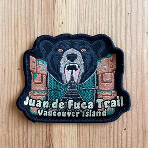 May include: Embroidered patch featuring a black bear with a snarling expression, flanked by totem-like structures. The text "Juan de Fuca Trail Vancouver Island" is displayed in white and green. The patch has a black border and is set against a wooden background.