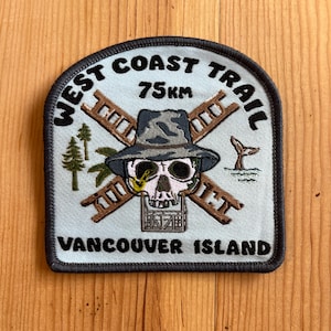May include: Embroidered patch with a light blue background. It features a skull wearing a hat, crossed ladders, and the text "WEST COAST TRAIL 75km VANCOUVER ISLAND". Includes a whale tail and trees.