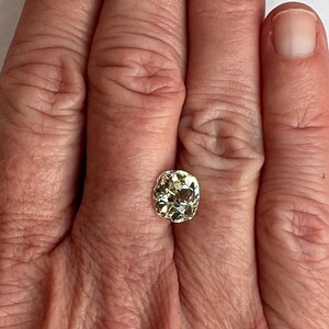 May include: A pale yellow oval-shaped gemstone with a faceted cut. The gemstone is set in a ring and is being worn on a person's finger.