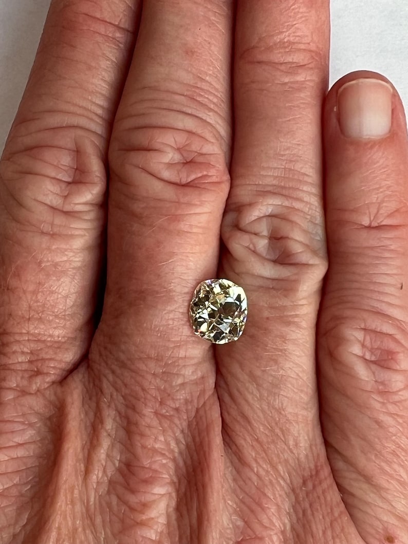 May include: A pale yellow oval-shaped gemstone with a faceted cut. The gemstone is set in a ring and is being worn on a person's finger.