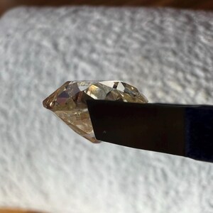 May include: A clear, faceted, light brown gemstone held in a black metal tweezer.