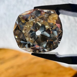 May include: A clear, faceted gemstone with a hole in the centre. The gemstone is held by a pair of tweezers and has a brown, amber-like hue.
