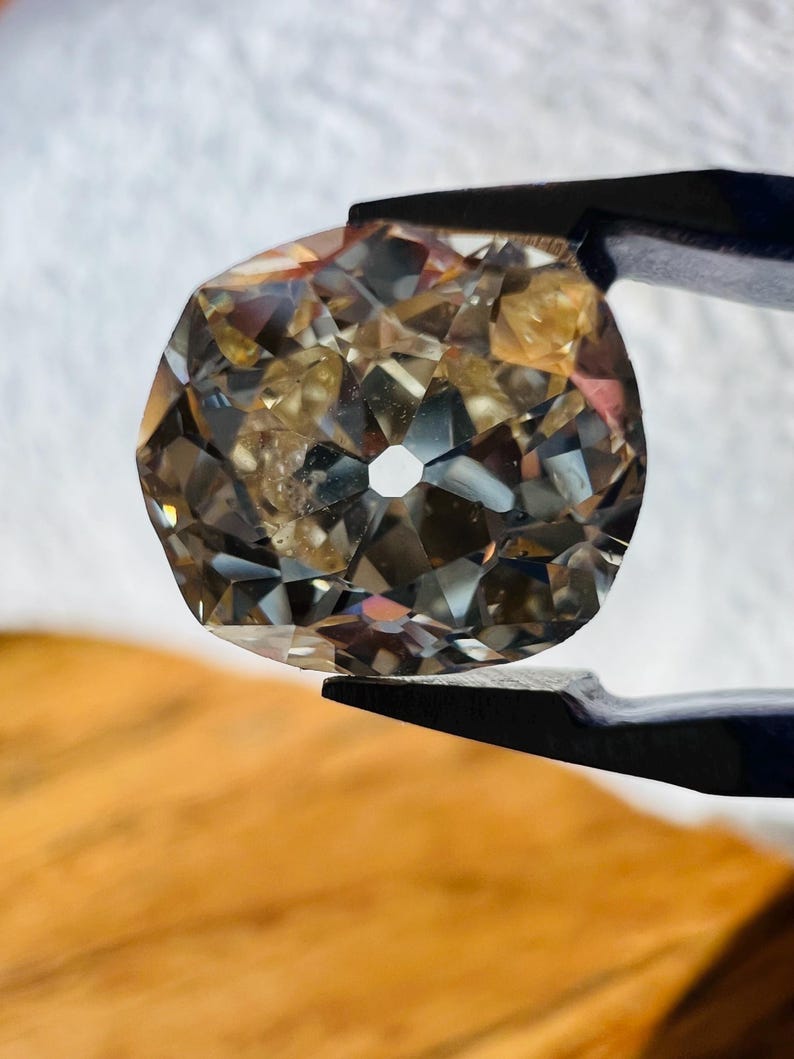 May include: A clear, faceted gemstone with a hole in the centre. The gemstone is held by a pair of tweezers and has a brown, amber-like hue.