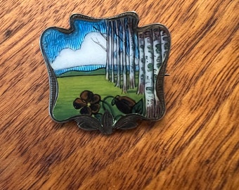 May include: A silver brooch with a blue and green enamel design depicting a forest scene with trees and a cloud. The brooch has a detailed floral design around the edge.