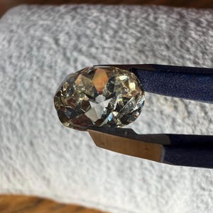 May include: A clear, faceted, oval-shaped gemstone held by tweezers. The gemstone has a light brown hue and reflects light.