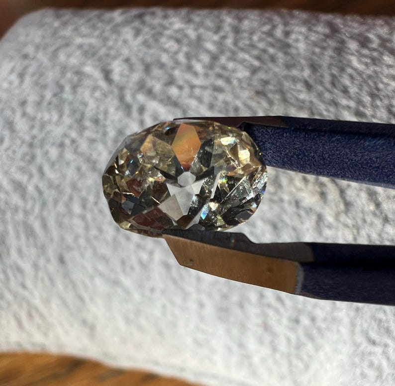 May include: A clear, faceted, oval-shaped gemstone held by tweezers. The gemstone has a light brown hue and reflects light.