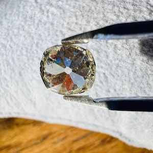 May include: A clear, faceted, cushion-shaped gemstone held by tweezers. The gemstone has a slightly yellowish hue.