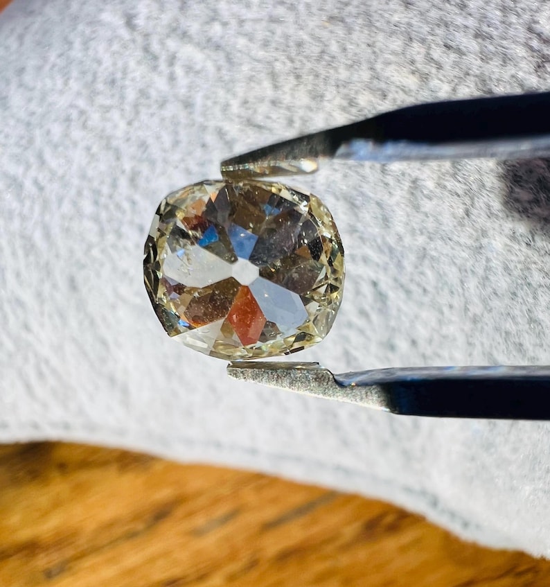 May include: A clear, faceted, cushion-shaped gemstone held by tweezers. The gemstone has a slightly yellowish hue.