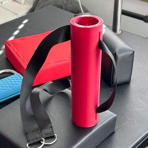 Pode incluir: Uma almofada cilíndrica de espuma rosa para barra de pesos com uma ranhura vertical ao longo do comprimento e uma correia de nylon preta fixada nas laterais. A correia inclui dois anéis em D de metal prateado na extremidade para fixação.