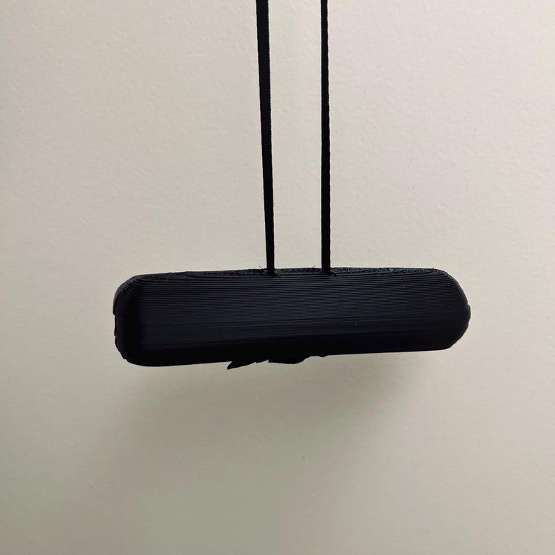May include: A black, rectangular object with rounded edges is suspended by two black cords. The object appears to be 3D-printed, with visible layer lines. The background is a plain, off-white wall.