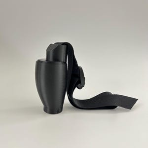 Puede incluir: Un objeto negro impreso en 3D con una base redondeada y una parte superior rectangular. Una correa negra con una hebilla está unida al objeto. El objeto parece ser un tipo de soporte o montaje.