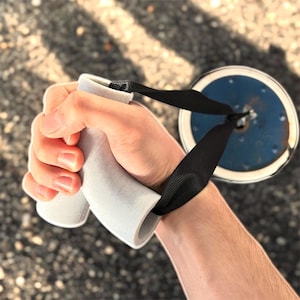 May include: A gray weightlifting handle with a black strap is held in a hand. The handle is connected to a blue and silver weight plate. The handle is cylindrical and has a textured surface.