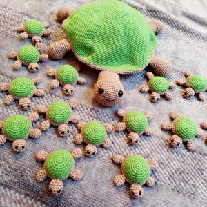 Könnte beinhalten: Eine Sammlung gehäkelter grüner und brauner Schildkrötenspielzeuge. Die Schildkröten sind in verschiedenen Größen und auf einer grauen, strukturierten Oberfläche angeordnet.