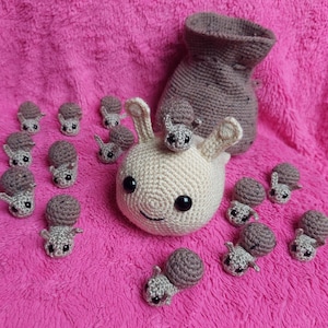 Puede incluir: Colección de artículos de ganchillo hechos a mano sobre un fondo rosa. El conjunto incluye un caracol grande, de color crema, con un caracol más pequeño encima, una bolsa marrón con cordón y varios caracoles grises pequeños. Los caracoles tienen ojos negros.
