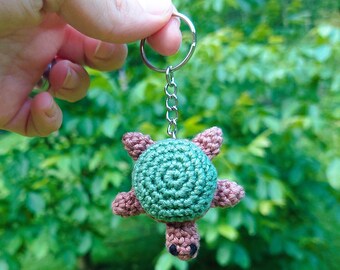 May include: A handmade crochet turtle keychain. The turtle has a green shell, brown flippers, and a brown head with black eyes. The keychain is attached to a silver metal ring and chain.