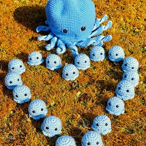 Puede incluir: Un pulpo de crochet azul rodeado por una forma de corazón de pulpos de crochet azules más pequeños sobre un lecho de hierba marrón.