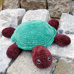 Puede incluir: Un peluche de tortuga con caparazón turquesa y extremidades color burdeos. La tortuga tiene un caparazón redondo y texturizado, y las extremidades son ovaladas. Tiene dos ojos pequeños, redondos y morados. El juguete está hecho de un material suave y difuso.