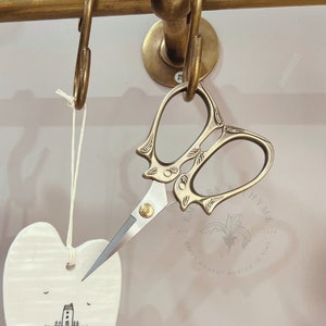May include: White ceramic heart-shaped ornament with the text "Love makes a house into a home" and a small illustration of a house. The ornament is hanging from a hook with a pair of gold-toned scissors.
