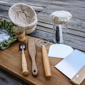 May include: A collection of bread-making tools on a wooden surface. Includes a proofing basket, a jar with a cloth cover, a dough scraper, a lame, a wooden spoon, a dough whisk, and a bench scraper. The tools are made of wood, metal, and fabric.