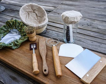 May include: A collection of bread-making tools on a wooden surface. Includes a proofing basket, a jar with a cloth cover, a dough scraper, a lame, a wooden spoon, a dough whisk, and a bench scraper. The tools are made of wood, metal, and fabric.
