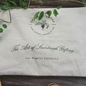 May include: Cream-colored canvas bag with the text "The Art of Sourdough Gifting" and "Los Angeles California" printed in dark green. The bag also features a circular logo with text and a botanical illustration. Green leaves are draped over the bag.