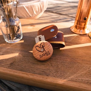 May include: A wooden shaving soap puck with the text "ISAIAH 41:10" and a heart and cross design, alongside a leather razor case and a straight razor blade. The items are on a wooden surface with copper-colored items in the background.