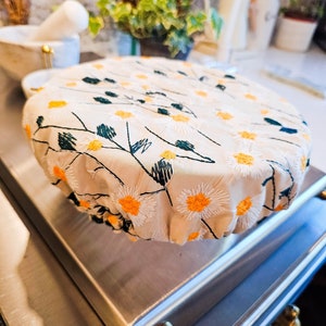 May include: A beige fabric food cover with a floral pattern of white daisies with yellow centers and green stems. The cover is on top of a stainless steel surface.