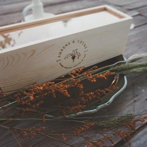 May include: A light-colored wooden box with a clear lid, featuring the text "EMBERS & THYME" and a floral design. The box is resting on a silver-colored tray, with dried orange and green foliage.