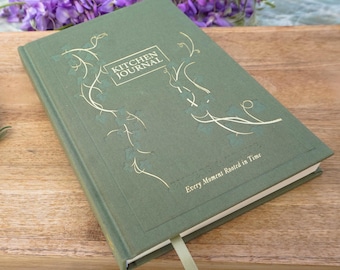 May include: A green hardcover journal with gold lettering that reads "Kitchen Journal" and the text "Every Moment Rooted in Time". The journal has a decorative vine design on the cover.