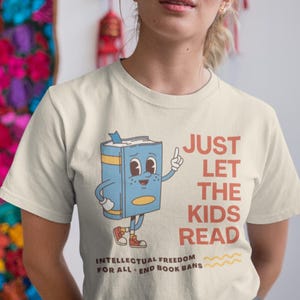 May include: Cream-colored t-shirt with the text "JUST LET THE KIDS READ" in red. A cartoon book character is pointing upwards. The shirt also has the text "INTELLECTUAL FREEDOM FOR ALL - END BOOK BANS".