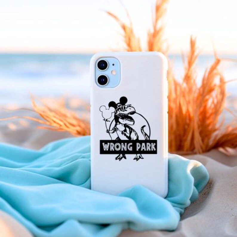 May include: White phone case with a black graphic of a dinosaur wearing sunglasses and holding a Mickey Mouse balloon. The text "WRONG PARK" is below the graphic. The phone case is on a sandy surface with blue fabric.