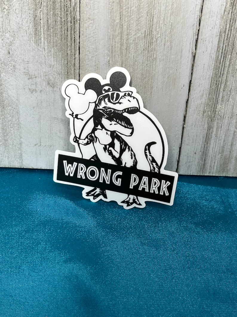 May include: A black and white sticker featuring a dinosaur wearing sunglasses and holding a Mickey Mouse balloon. The text "WRONG PARK" is displayed on a black rectangle below the dinosaur.