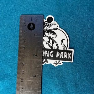 May include: A black and white sticker featuring a dinosaur wearing sunglasses and a Mickey Mouse-style head. The sticker reads "ONG PARK" in a stylized font. The sticker measures approximately 4 inches in height.