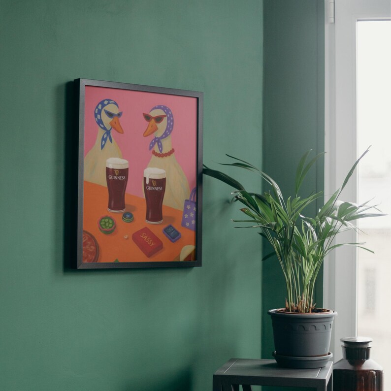 K&ouml;nnte beinhalten: Gerahmter Kunstdruck mit zwei G&auml;nsen mit Sonnenbrillen und Kopft&uuml;chern, jede mit einem Pint Guinness. Das Kunstwerk hat einen rosa Hintergrund und einen orangen Tisch mit dem Wort "SASSY" auf einer roten Karte.