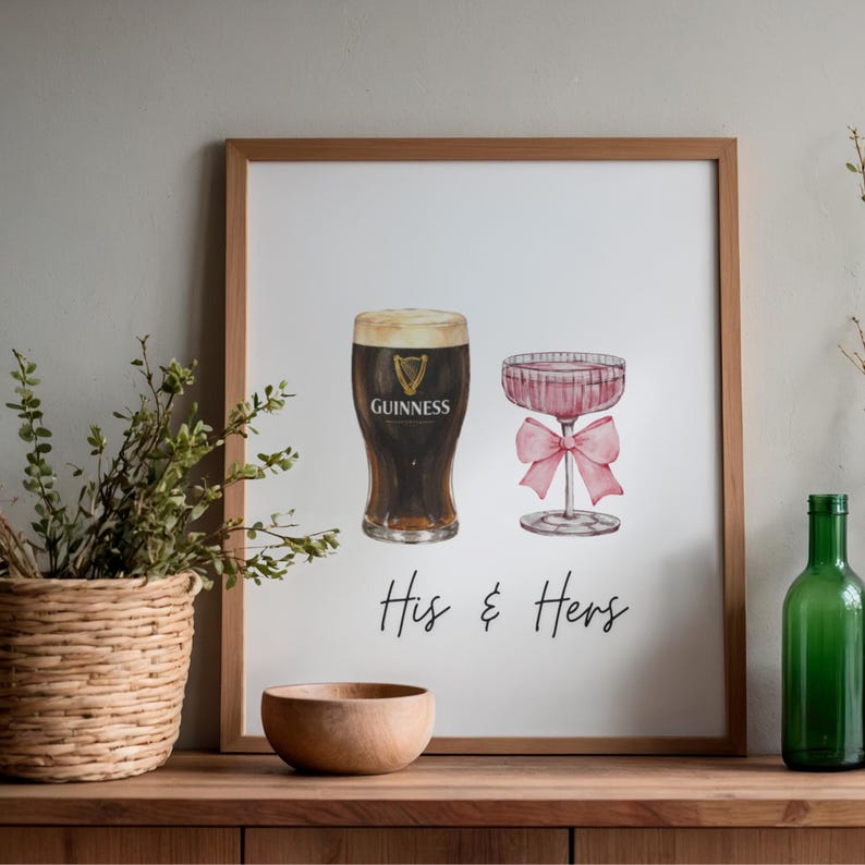 Puede incluir: Una impresi&oacute;n enmarcada con ilustraciones de acuarela de una pinta de Guinness y una copa de champ&aacute;n rosa. El texto "His & Hers" est&aacute; escrito debajo. La impresi&oacute;n se muestra sobre una superficie de madera.