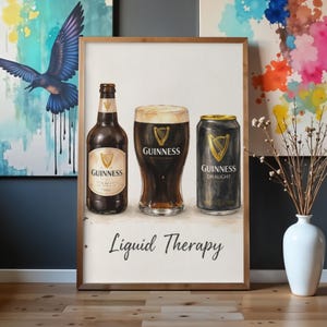 May include: A framed print depicting a bottle, a pint glass, and a can of Guinness. The print includes the words "Liquid Therapy". The Guinness logo is visible on each item. The background is white, with a wooden frame.