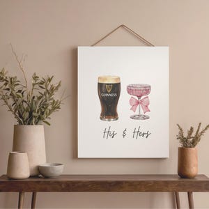 Puede incluir: Impresi&oacute;n enmarcada con ilustraciones de acuarela de una pinta de Guinness y una copa de champ&aacute;n con un lazo rosa. Las palabras "His & Hers" est&aacute;n escritas debajo. La impresi&oacute;n est&aacute; colgada en una pared.