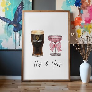 Puede incluir: Una impresi&oacute;n enmarcada con ilustraciones de acuarela de una pinta de Guinness y una copa de champ&aacute;n rosa con un lazo. Las palabras "His & Hers" est&aacute;n escritas debajo. La obra de arte se exhibe sobre una superficie de madera.