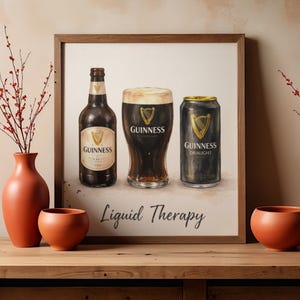 May include: A framed print depicting a bottle, a pint glass, and a can of Guinness. The words "Liquid Therapy" are at the bottom. The artwork is displayed on a wooden surface with orange vases and a plant.