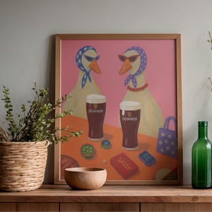K&ouml;nnte beinhalten: Ein gerahmter Kunstdruck mit zwei G&auml;nsen mit Sonnenbrillen und Kopft&uuml;chern, mit zwei Pints Guinness. Das Kunstwerk hat einen rosa Hintergrund und einen Tisch mit einer Holzschale, einer roten Schachtel mit der Aufschrift "SASSY" und einer kleinen Handtasche.