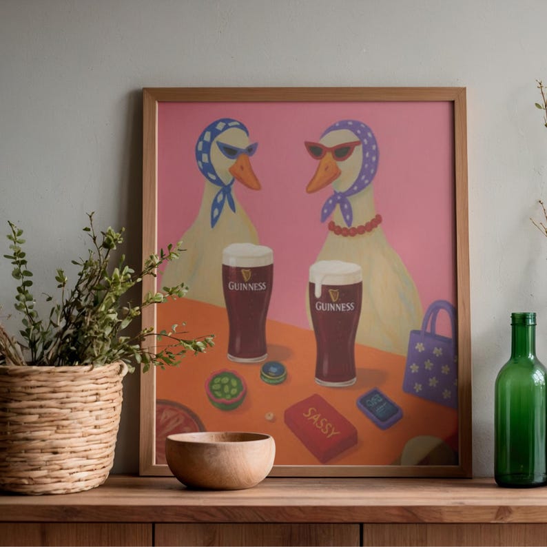 K&ouml;nnte beinhalten: Ein gerahmter Kunstdruck mit zwei G&auml;nsen mit Sonnenbrillen und Kopft&uuml;chern, mit zwei Pints Guinness. Das Kunstwerk hat einen rosa Hintergrund und einen Tisch mit einer Holzschale, einer roten Schachtel mit der Aufschrift "SASSY" und einer kleinen Handtasche.