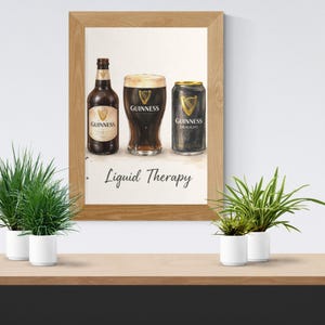 May include: A framed print showing a watercolour illustration of Guinness stout. The image includes a bottle, a pint glass, and a can of Guinness, with the text "Liquid Therapy" below. The frame is light brown.