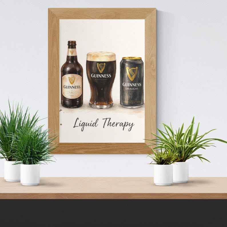 May include: A framed print showing a watercolour illustration of Guinness stout. The image includes a bottle, a pint glass, and a can of Guinness, with the text "Liquid Therapy" below. The frame is light brown.
